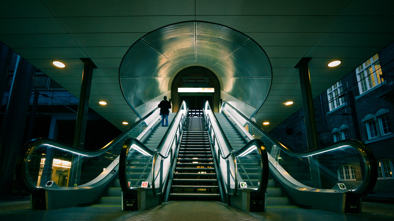 Two spacey looking escalators pointing up to the entrance of the station, one person on the left escalator going up.