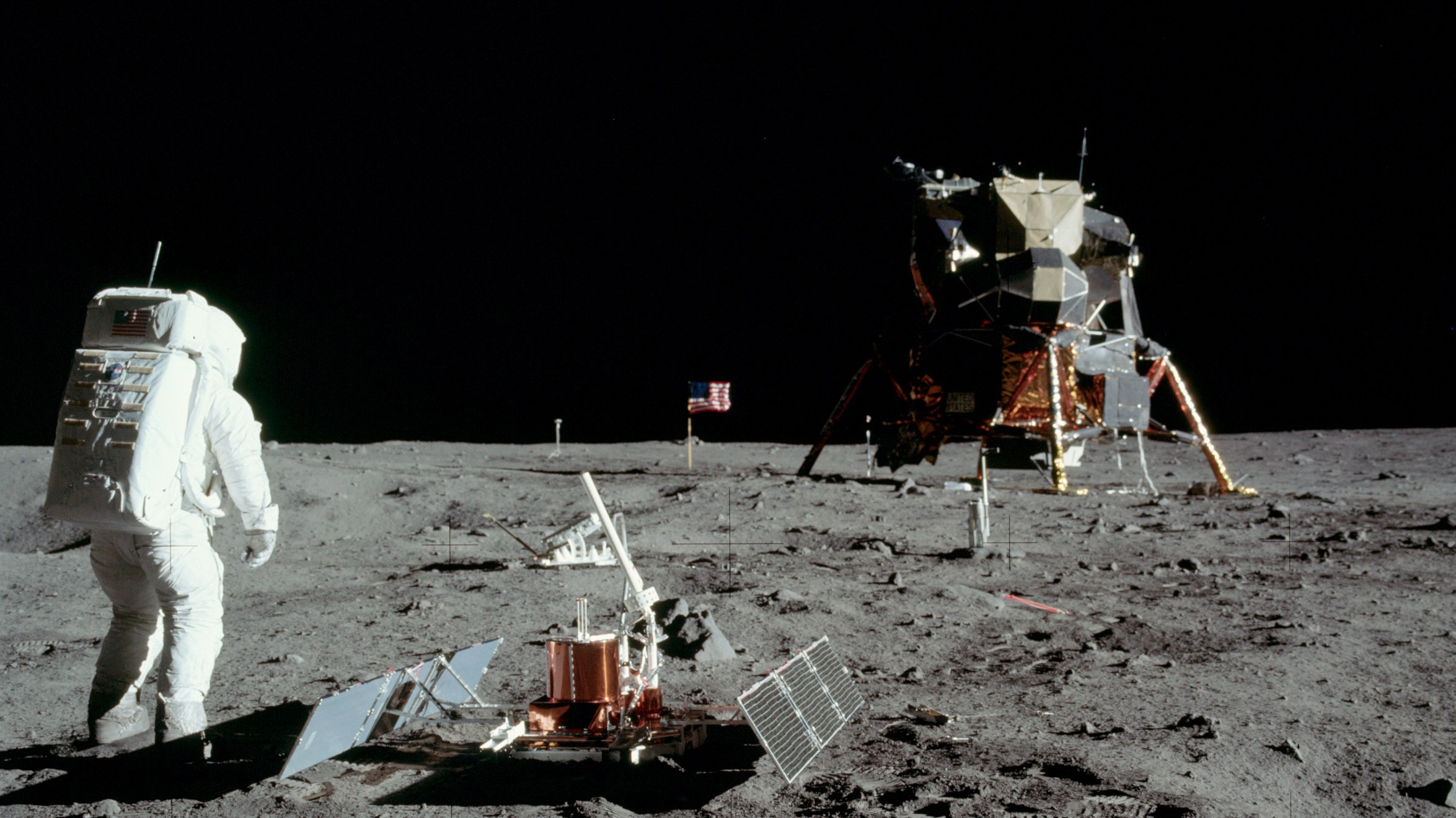 Buzz Aldrin on the moon with Landing Capsule in the background and technology experiment in the front.